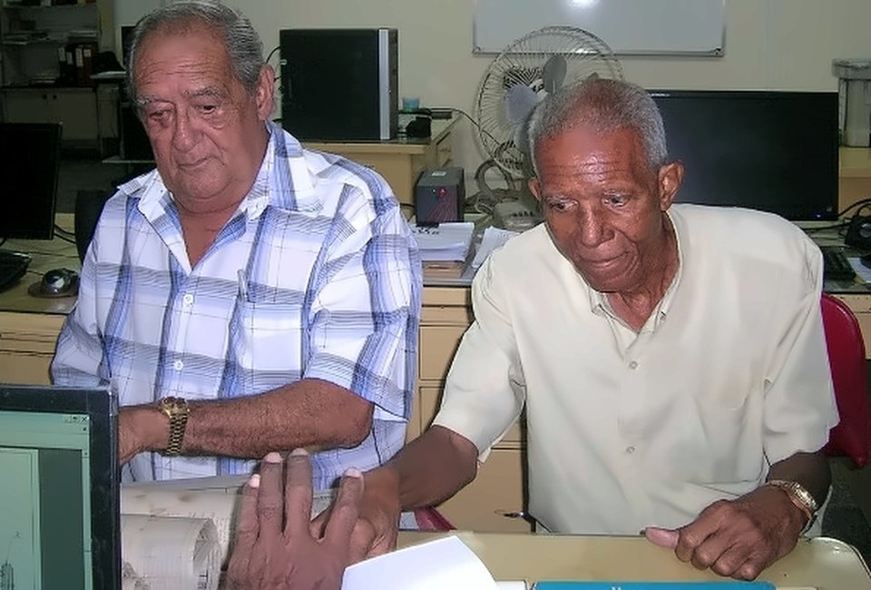 Dos hombres mayores trabajan en una oficina técnica, sentados frente a planos y computadoras.